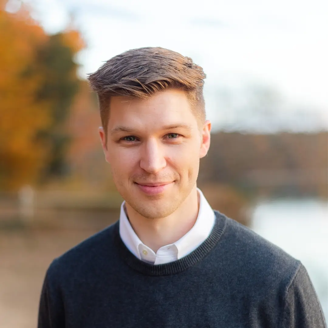 ben smiling while wearing a dark gray shirt with a white button undershirt in front of a blurred nature background
