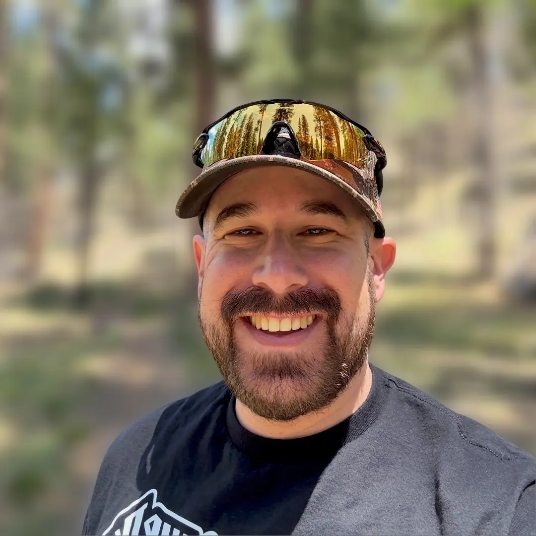 matt smiling while wearing a camo baseball cap and black shirt while hiking a dirt path surrounded by trees